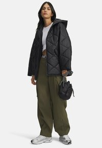 Černá prošívaná péřová bunda s kapucí, zelené široké cargo kalhoty, stříbrné tenisky a malá černá taška se šňůrkovým uzávěrem.