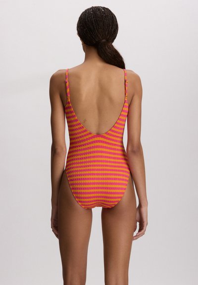 Γυναίκα με πλεξούδες, φορώντας μαγιό με χαμηλή πλάτη και υφή, με οριζόντιες πορτοκαλί και ροζ ρίγες, στέκεται μπροστά σε απλό φωτεινό φόντο.