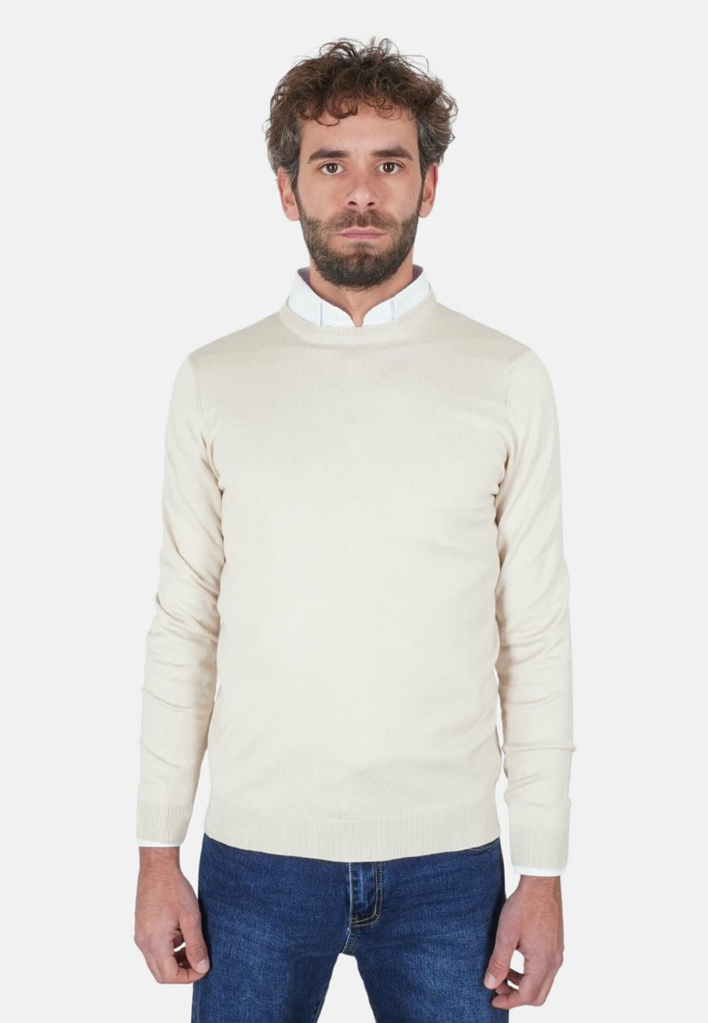 Uomo con capelli ricci e barba che indossa una camicia bianca con colletto, sopra una maglia crema e jeans blu, in piedi davanti a uno sfondo bianco.