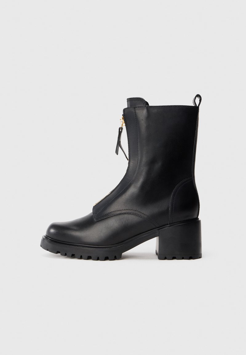 Bota de tornozelo em couro preto com salto robusto e fecho éclair frontal, apresentando uma alça de puxar e sola portada rugged sobre um fundo simples.