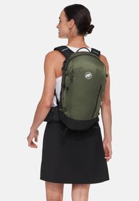 Zaino verde oliva con dettagli neri, compartimenti con zip, spallacci regolabili e un logo sulla parte anteriore. Indossato da una persona.
