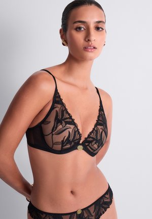 Femme portant une lingerie en dentelle noire transparente avec des motifs floraux et des accents dorés, debout sur un fond gris clair uni.