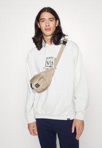 Bolso cruzado beige con forma curvada y tejido texturizado, que cuenta con un compartimento con cremallera y una correa ajustable, llevado sobre una sudadera blanca.