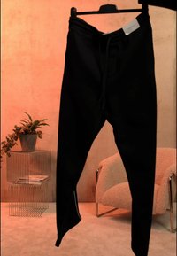 Črne ozke trenirke s vrvico na obešalniku, postavljene v notranjosti blizu žične police s rastlino in bež oblazinjeno stolom.