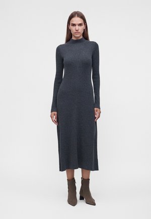 Femme portant une robe midi en maille côtelée gris foncé à manches longues et des bottines marron, debout devant un fond clair uni.