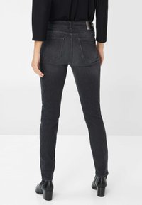 Personne portant un jean skinny noir et des bottines à talons noires, debout de dos, faisant face à l'appareil photo devant un fond uni.