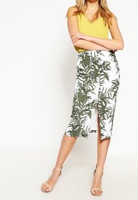 Vit kjol med grönt bladmönster, med en slits på sidan, kombinerad med en gul ärmlös topp och ljusbruna sandaler med remmar.