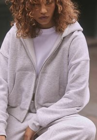 Felpa grigia con zip realizzata in tessuto morbido, caratterizzata da una vestibilità rilassata, ampie tasche e polsini a coste, indossata sopra una maglietta bianca.