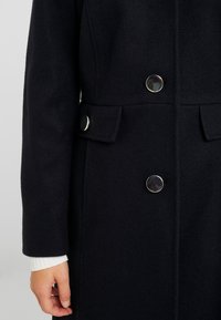 Cappotto di lana nera con grandi bottoni argentati sulla parte anteriore e tasca a pattina, con polsino di maglione bianco a coste visibile al polso.