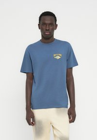Μπλε βαμβακερό t-shirt με κίτρινο λογότυπο Rip Curl στο στήθος. Κοντά μανίκια και κλασικός στρογγυλός γιακάς. Ομαλή υφή.