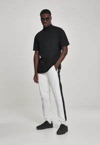 Svart kortermet t-skjorte med høy krage, kombinert med hvite bukser med svarte sidestriper, og svarte sneakers, på en ensfarget bakgrunn.