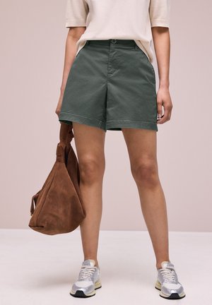 Osoba ubrana w ciemnozielone szorty, biały krótki rękaw, srebrne trampki, trzyma dużą brązową zamszową torbę przy boku.
