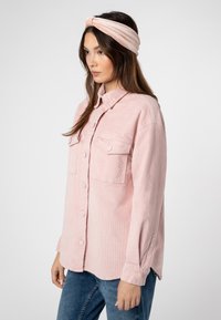 Roze korduroy overhemd met een knoopsluiting aan de voorkant, twee voorzakken, lange mouwen en een kraag, gedragen met een bijpassende haarband.