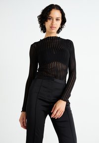 Schwarzes Langarmoberteil mit geripptem Muster und transparentem Streifendesign, das einen gewellten Ausschnitt hat. Kombiniert mit einer hochgeschnittenen schwarzen Hose.
