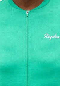 Κοντινή λήψη σε ποδηλατική φανέλα μπλε-πράσινου χρώματος με υφή, με μπροστινό φερμουάρ και λευκό λογότυπο «Rapha» στο αριστερό στήθος.