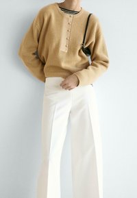 Beżowy prążkowany sweter z okrągłym dekoltem i listwą na guziki, w połączeniu z białymi spodniami o szerokich nogawkach, o gładkiej teksturze.