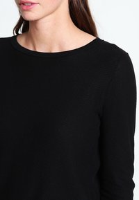 Kobieta z długimi brązowymi włosami, ubrana w czarną, teksturowaną bluzkę z długim rękawem i okrągłym dekoltem, na białym tle.