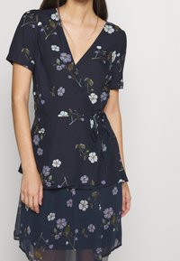 Haut à imprimé floral en tissu bleu marine foncé avec un décolleté en V, manches courtes et taille nouée, présentant des fleurs blanches et violettes ainsi que des feuilles vertes.