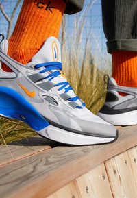 Graue Sneakers mit blauen Schnürsenkeln und orangen Akzenten. Verfügen über ein Obermaterial aus Netzstoff, eine gepolsterte Sohle und ein geschwungenes Design. Getragen mit leuchtend orangenen Socken.