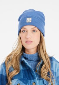 Cappello lavorato a maglia blu con risvolto, caratterizzato da una toppa quadrata con un logo di montagna. Indossato con un dolcevita a costine blu chiaro.