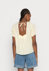 Blusa amarela de mangas curtas com padrão xadrez, costas abertas com laço e bainha franzida. Usada com calções de ganga com punhos dobrados.