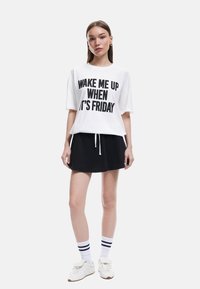 Hvid bomulds t-shirt med sort tekst "VÆK MIG NÅR DET ER FREDAG," parret med en sort jersey-nederdel. Hvide sko og stribede sokker fuldender looket.