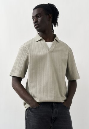 Fiatal fekete férfi dreadlockkal, bézs rövid ujjú galléros ingben és fekete farmerral, kezét a zsebében tartva, oldalra néz egy fehér háttér előtt.