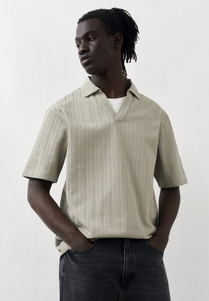 Νέος μαύρος άνδρας με dreadlocks που φοράει μπεζ κοντομάνικο πουκάμισο με γιακά και μαύρα τζιν, με τα χέρια στις τσέπες, κοιτάζοντας πλάγια πάνω σε λευκό φόντο.