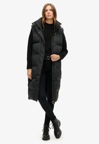 Schwarze puffy lange Weste mit Kapuze, ausgestattet mit einem Reißverschluss vorne und Druckknöpfen, kombiniert mit einem schwarzen Pullover und engen Hosen.