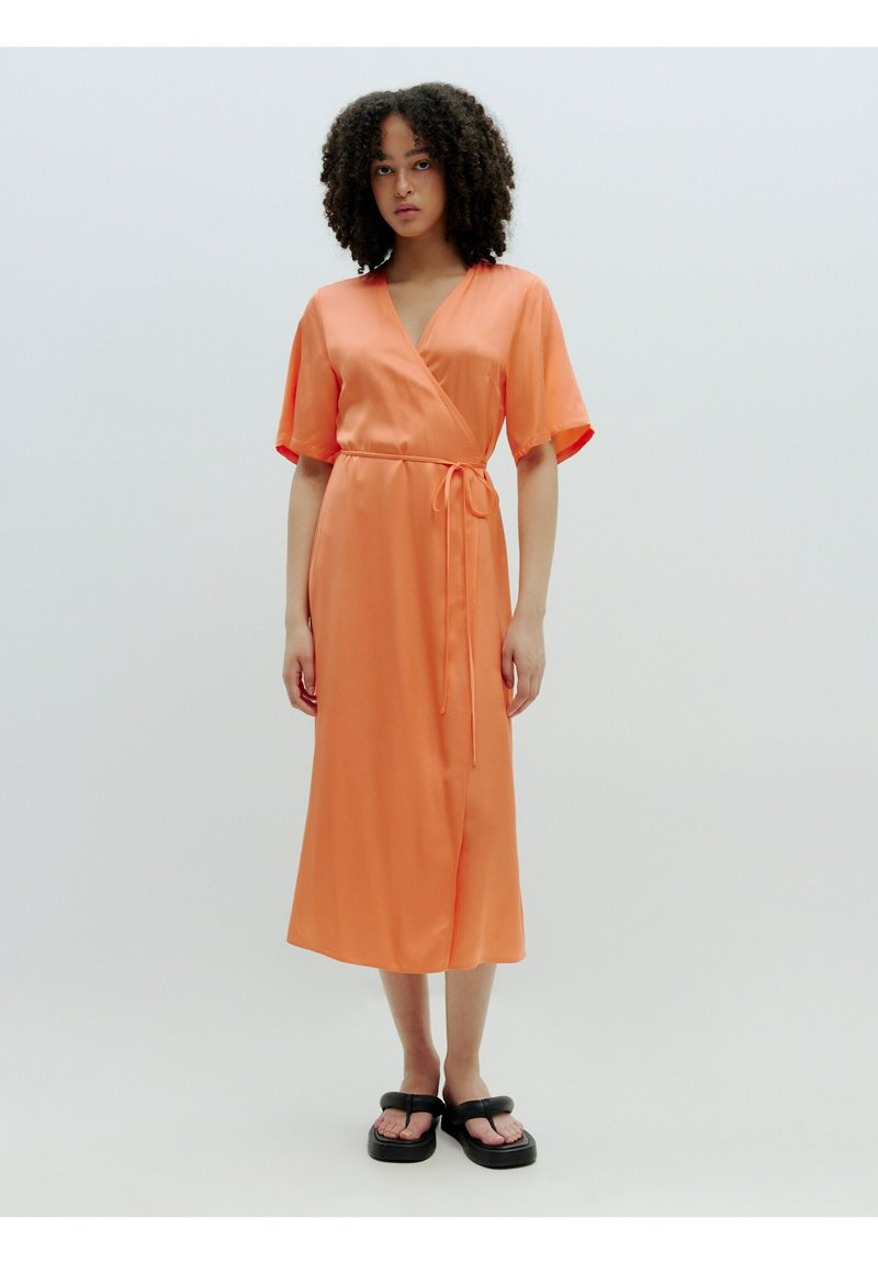 Oranje satijnen omslagjurk met korte mouwen en een strik taille. Heeft een middellange snit en een gladde, glanzende textuur. Gepaard met zwarte sandalen.