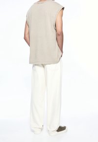 Sleeveless beige cotton shirt with a relaxed fit, paired with loose white pants and olive green sneakers. Simple design with side pockets.