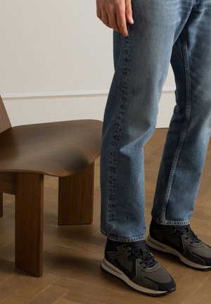 Lichte blauwe denim jeans, gecombineerd met zwarte en grijze sneakers. Een houten stoel in de buurt heeft een golvende zitting en vier stevige poten.