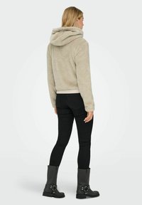 Woman facing away wearing beige fuzzy hooded jacket, black skinny jeans, and black mid-calf boots with buckle details.