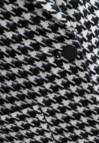 Svartvitt tygliknande mönster med hundtandsmönster och luddig textur; har en rund svart knapp och en synlig söm på fickan.