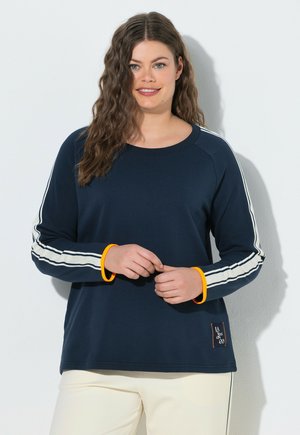 Kvinde med langt bølget hår iført en marineblå langærmet top med hvide striber og orange manchetter, smilende, stående mod en lys baggrund.