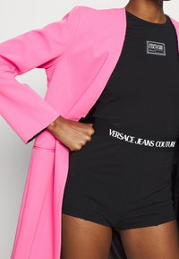 Ljusrosa skräddarsydd kavaj med långa ärmar, kombinerad med en svart bodysuit med en logoband. Slät textur, modern design.