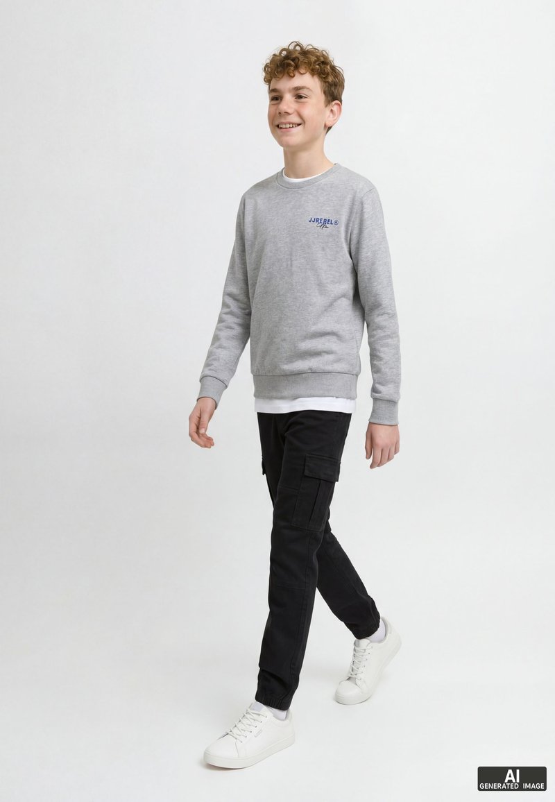 Adolescent marchant, portant un sweat-shirt gris clair, un pantalon cargo noir et des baskets blanches, souriant et regardant devant.