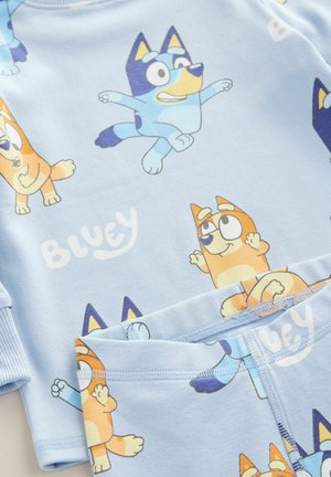 Lyseblå pyjamaser med tegneseriehunde i forskellige stillinger, i en orange og blå farveskema. "BLUEY"-tekst i et legende skrifttype. Blød stof.