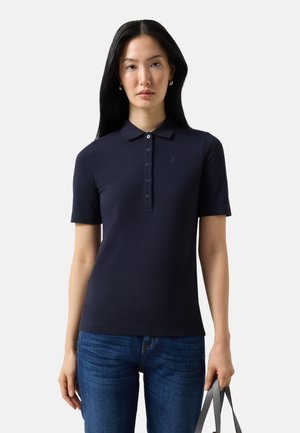 Kobieta o długich czarnych włosach, ubrana w granatową koszulkę polo z krótkim rękawem i niebieskie jeansy, trzymająca szare paski torby w prawej ręce.