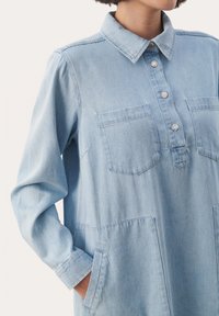 Vestito camicia in denim azzurro chiaro con colletto, chiusura a bottoni, due tasche sul petto e maniche lunghe con polsini. Design strutturato e casual.