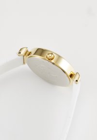 Boîtier de montre rond en or avec un bracelet en cuir texturé blanc. Présente une couronne proéminente et un dos lisse et uni.