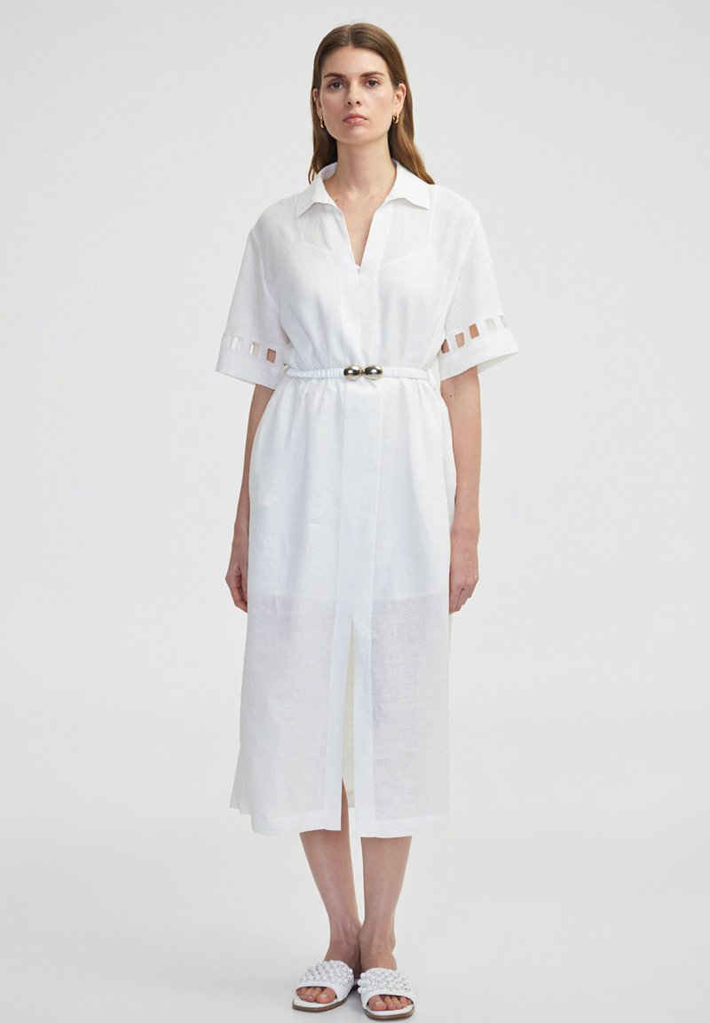Witte linnen jurk met een kraaghalslijn, korte mouwen en uitsnijdingen. Beschikt over een tailleriem met metallic accenten.