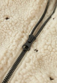 Tissu en polaire de couleur crème avec une surface texturée, doté d'une fermeture éclair noire et de minimalistes accents bruns près de la ligne de fermeture.