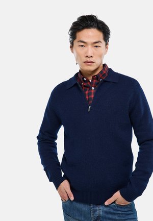 Jeune homme aux cheveux noirs portant un pull zippé bleu marine par-dessus une chemise à carreaux rouge et noir, les mains dans les poches, sur un fond blanc.