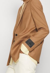Brauner Wollblazer mit taillierter Passform, zwei schwarzen Knöpfen, schwarzem Logo-Patch am Ärmel und einer geschichteten hellgelben Bluse darunter.