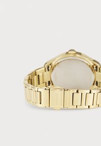 Montre en métal doré avec un bracelet à maillons, finition lisse et fermoir prononcé. La vue arrière montre un boîtier en acier inoxydable. Aucune caractéristique visible à l'arrière.