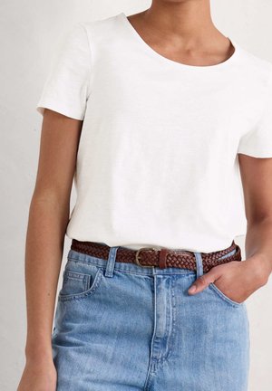 Person trägt ein weißes Kurzarmhemd, das in hoch taillierte blaue Jeans gesteckt ist, mit einem geflochtenen braunen Ledergürtel und einer Hand in der Tasche.