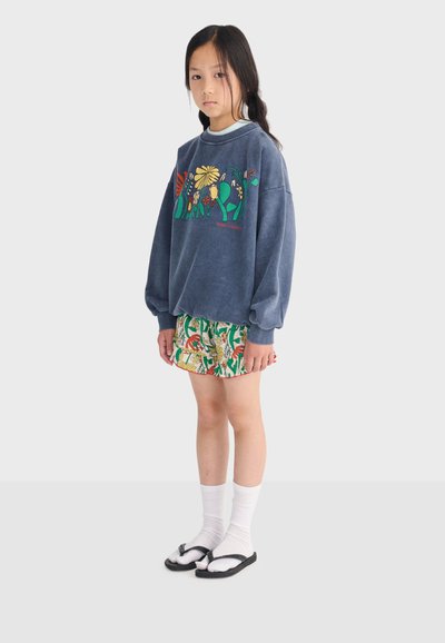 Jeune fille portant un sweat-shirt bleu à motifs floraux, un short à motifs, des chaussettes blanches et des tongs noires, posant devant un fond uni.