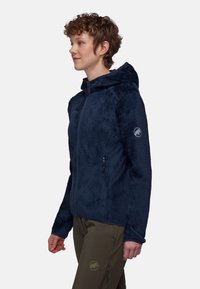 Marinblå huvjacka tillverkad av texturerad tyg, med dragkedjeslutning och dragkedjefickor på sidorna; logotyp på ärmen.