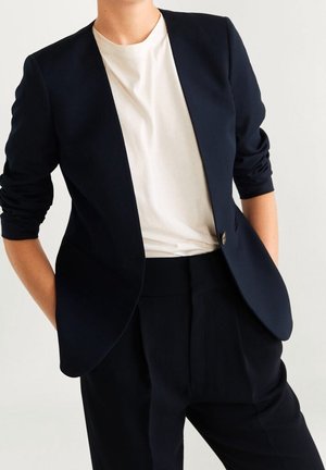 Osoba ubrana w dopasowaną czarną marynarkę z marszczonymi rękawami, pod którą ma białą koszulkę z okrągłym dekoltem, oraz czarne spodnie z wysokim stanem na jednolitym tle.